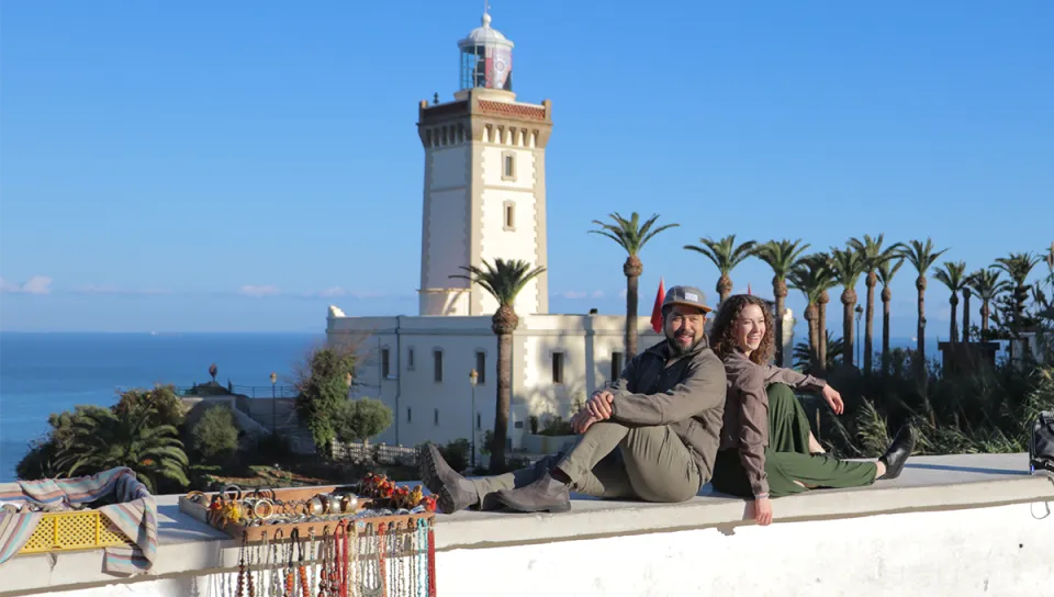 Two students pose with raised arms on a stone walkway at a medieval castle with a square tower visible behind them.