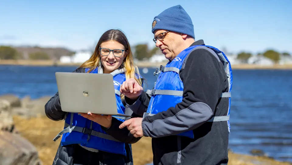 Student and instructor in blue life jackets examine a laptop together on a rocky shoreline with water and coastal landscape in the background.