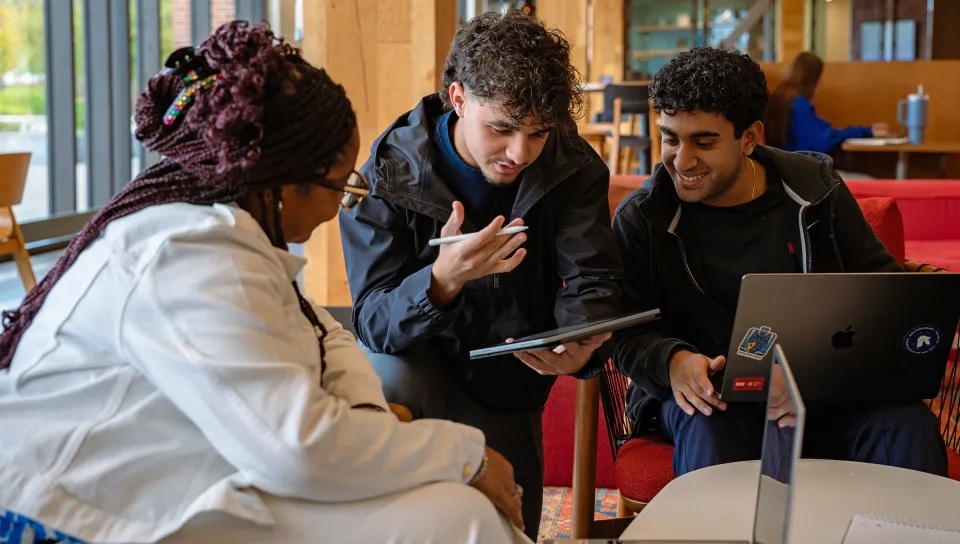 Three students collaborate in a study lounge, two working on laptops while the student in the center gestures while holding a tablet during their discussion.