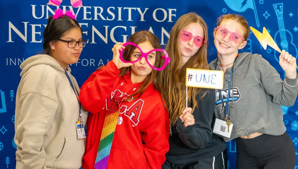 Four students pose with photo booth props, including oversized glasses, bunny ears, and a lightning bolt wand — in front of a University of New England backdrop, one holding a # U N E sign.