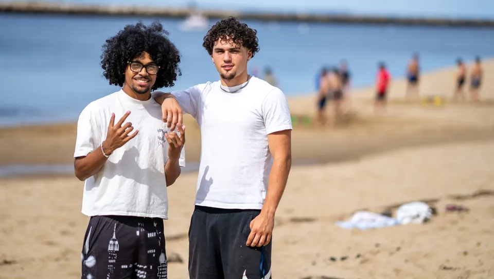 Two students pose together on a sandy beach with the water behind them.