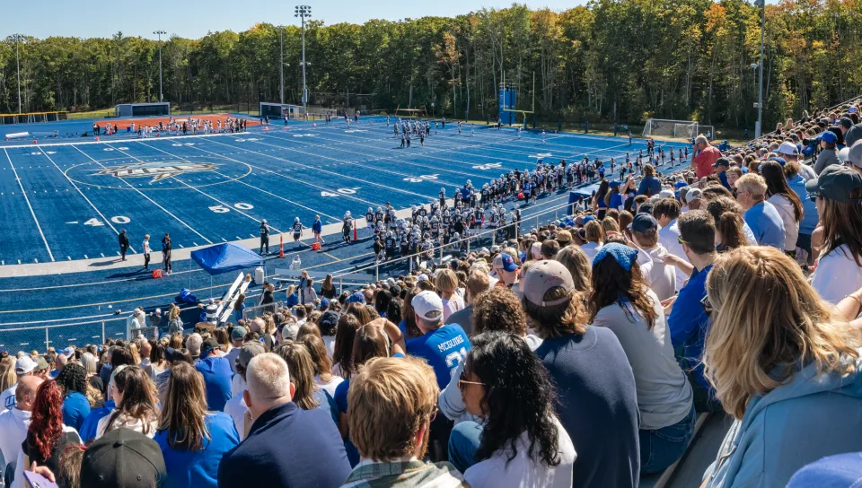 A packed crowd fills the stands overlooking U N E's blue turf football field during the 2025 Homecoming game, with players lined up along the sideline.