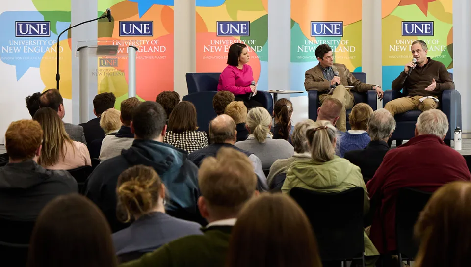 Three panelists speak into microphones on stage in front of a seated audience at a U N E President's Forum event, with U N E banners displayed behind them.