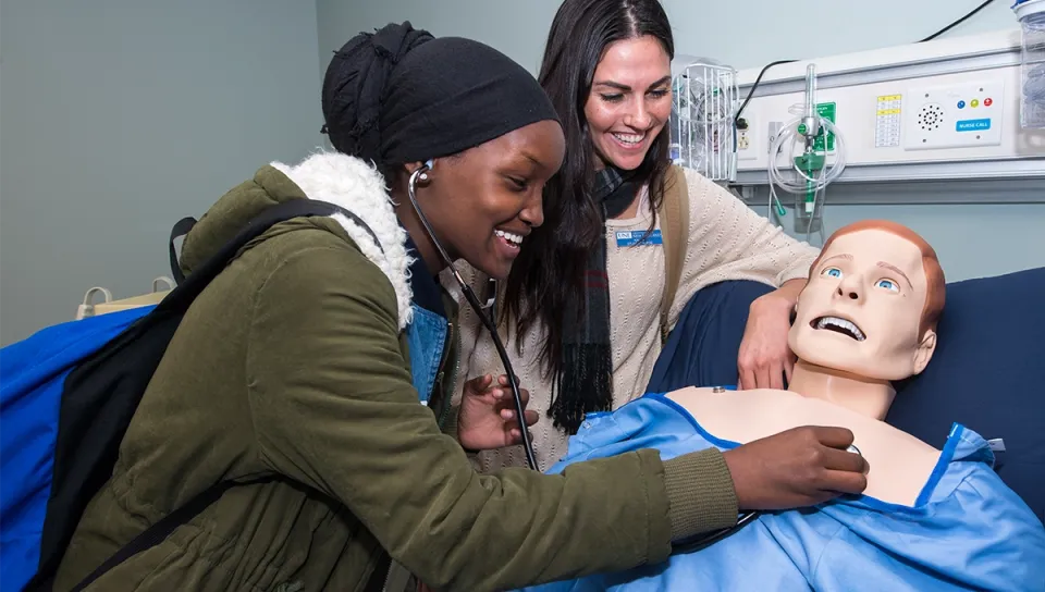A high school student uses a stethoscope on a medical mannequin while a U N E staff member guides her in a clinical simulation lab.