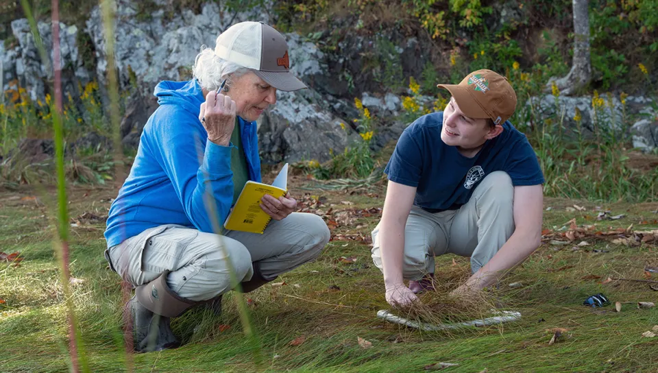 A U N E professor takes notes while a student examines grass within a sampling ring during outdoor fieldwork among coastal rocks.