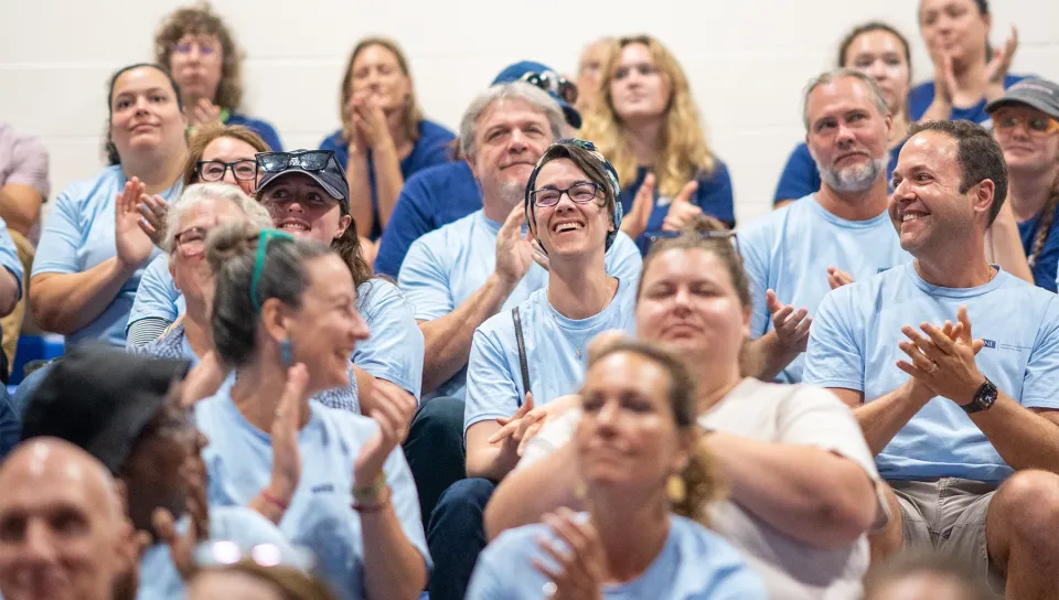 U N E employees in matching light blue T-shirts smile and applaud in the audience at the 2026 Fall Assembly.