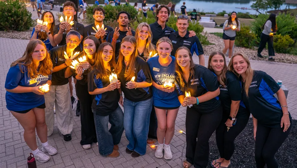 A large group of U N E student staff and orientation leaders pose together holding candles at the 2026 First Night event.