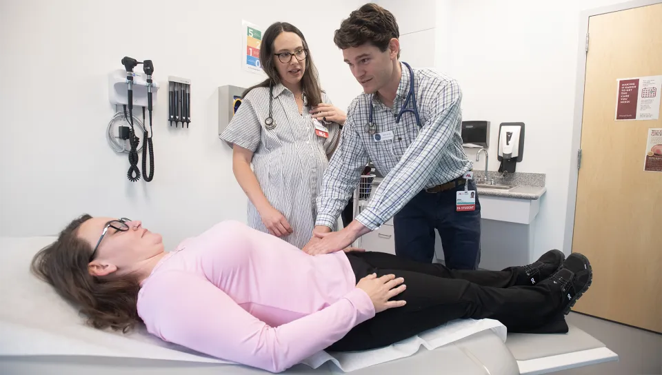 A U N E Physician Assistant student examines a patient's abdomen while a preceptor observes in a clinical exam room.
