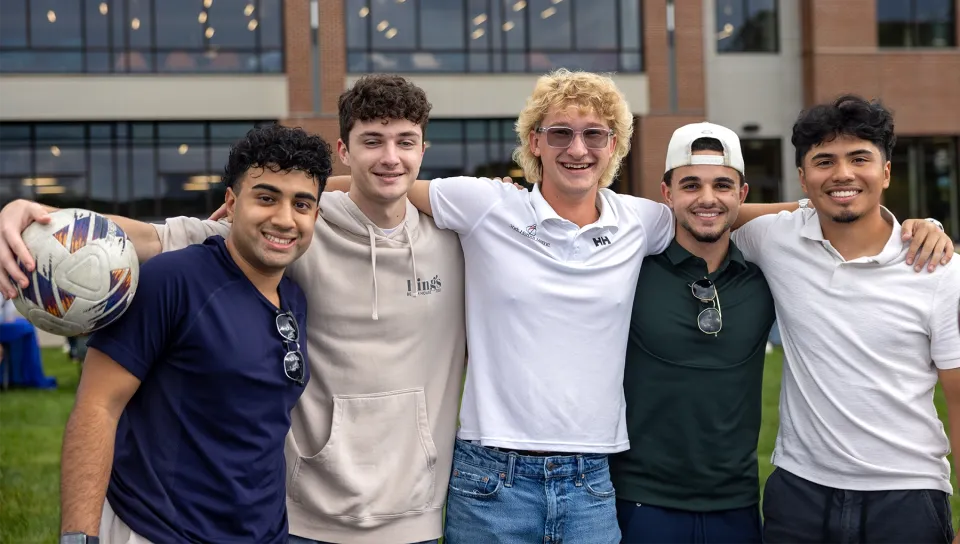 A group of five U N E students pose together with arms around each other at an outdoor welcome event.