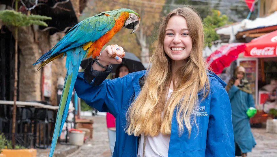 A U N E student in a university rain jacket smiles while holding a colorful macaw on a rainy street in Morocco.