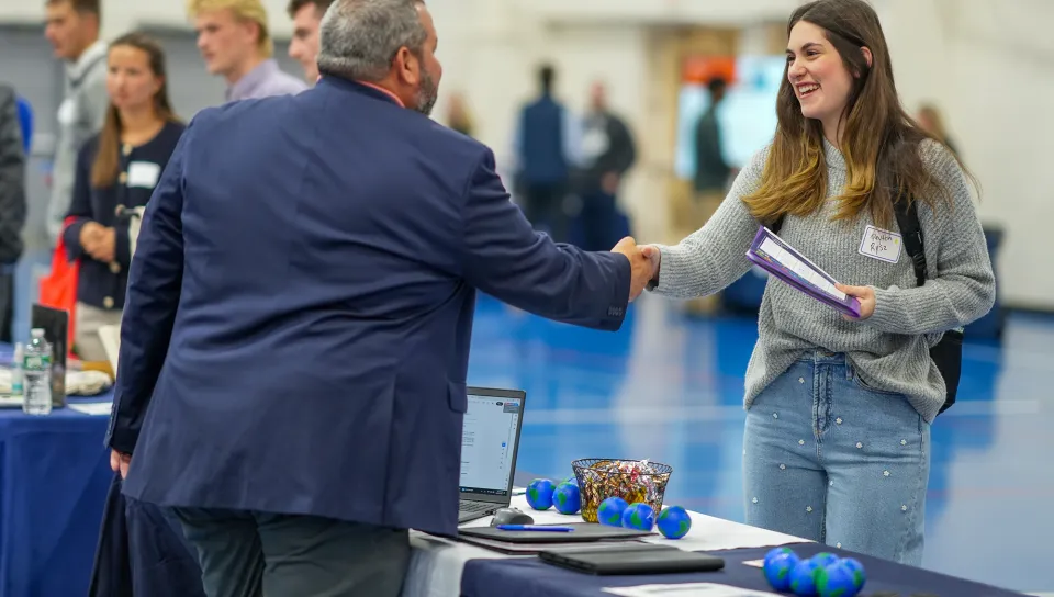 A smiling U N E student shakes hands with an employer at a booth during Industry Exploration Day.