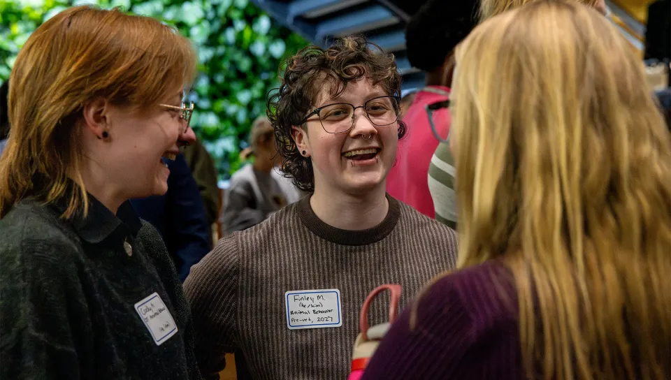 U N E students wearing name badges laugh together while chatting at an indoor mixer event.