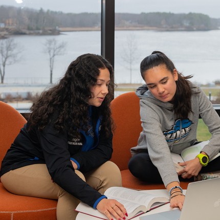 Two students studying in front of a large window in the Commons that overlooks the Saco River
