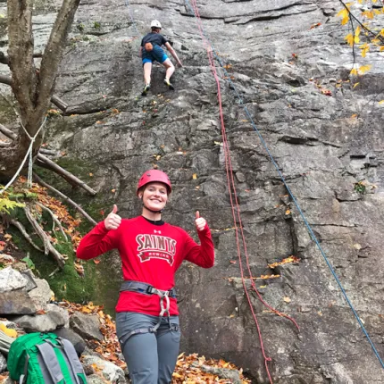 A student gives two thumbs up in front of a mountainside that another student is rock climbing