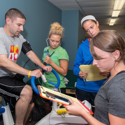 Three physical therapy students take notes as another student rides a stationary bike
