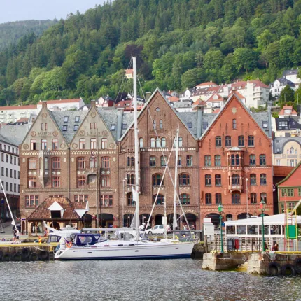 A boat drives through a river with brick buildings and mountains in the background