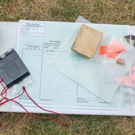 Archaeological field materials including a clipboard with shovel test record forms, brick fragments, and artifact bags used in the excavation of The Wayside site.