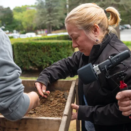 A student carefully examines soil and artifacts in a wooden screen during the Time Team New England archaeological fieldwork at UNE