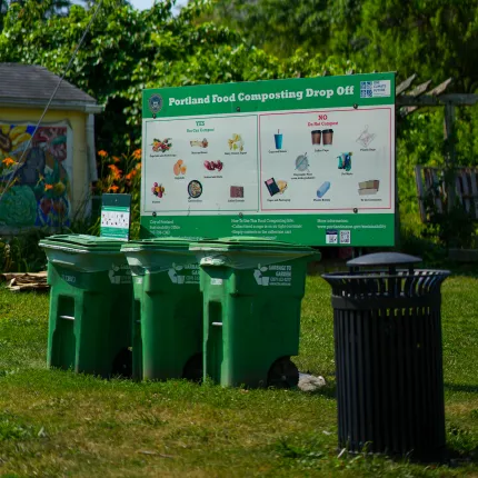 Portland food composting drop-off station with green collection bins and informational sign showing compostable and non-compostable items, set on grass with trees in background.