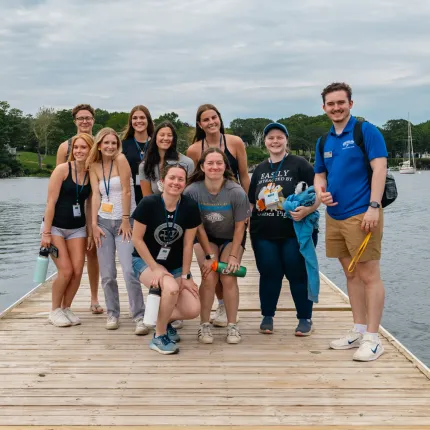 A group of U N E students and a tour guide pose together on a waterfront dock with sailboats visible in the harbor behind them.