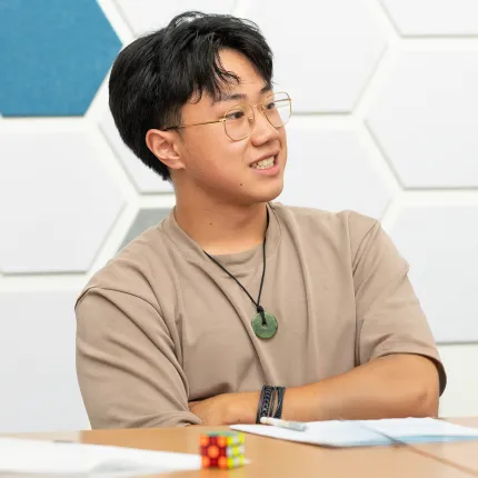 A student smiles and speaks during a discussion at a table in a modern common room. A geometric hexagonal wall panel installation in white and blue is visible in the background.