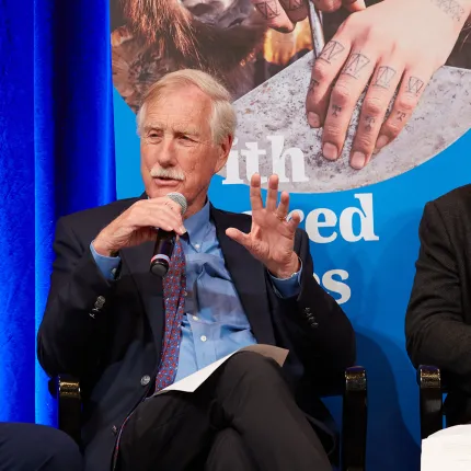 Angus King addresses an audience using a handheld microphone during a panel discussion, gesturing with one hand against a blue branded backdrop.