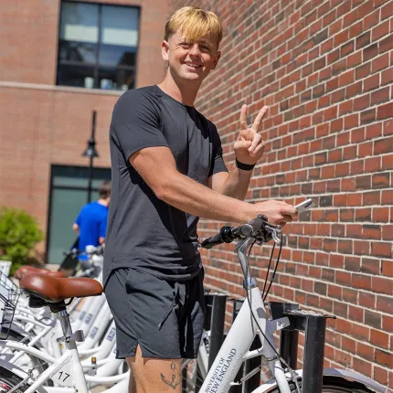 A smiling student flashes a peace sign while standing with a campus rental bike against a brick building wall.