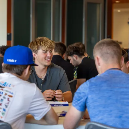 A smiling student chats with classmates seated around a table with a box of oil pastels in a busy campus commons area.