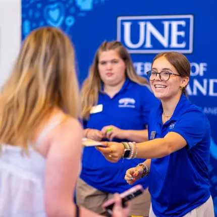 A smiling orientation staff member in a U N E polo hands paperwork to a student in front of a UNE banner.