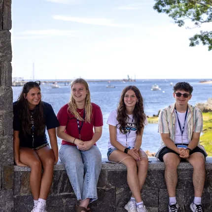 Four students in U N E apparel smile while sitting on a stone wall overlooking the Saco River.