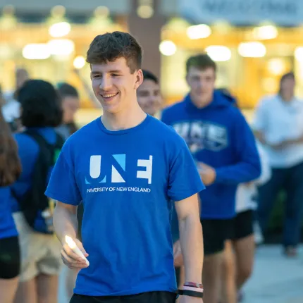 A smiling U N E student in a U N E T-shirt holds a candle during an evening First Night event on campus.
