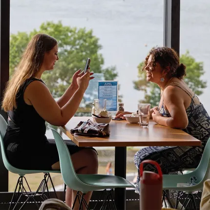 Two U N E students chat and share dessert at a table by the window in the Commons dining hall, overlooking the water.