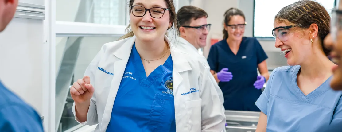 A student in a white coat talks with two other students in the anatomy lab