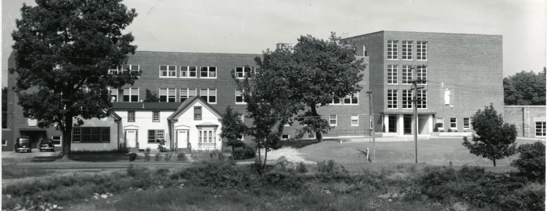 Historical black and white photograph showing Decary Hall at St. Francis College with the Wayside House, a small white house, in the foreground on UNE's Biddeford Campus.