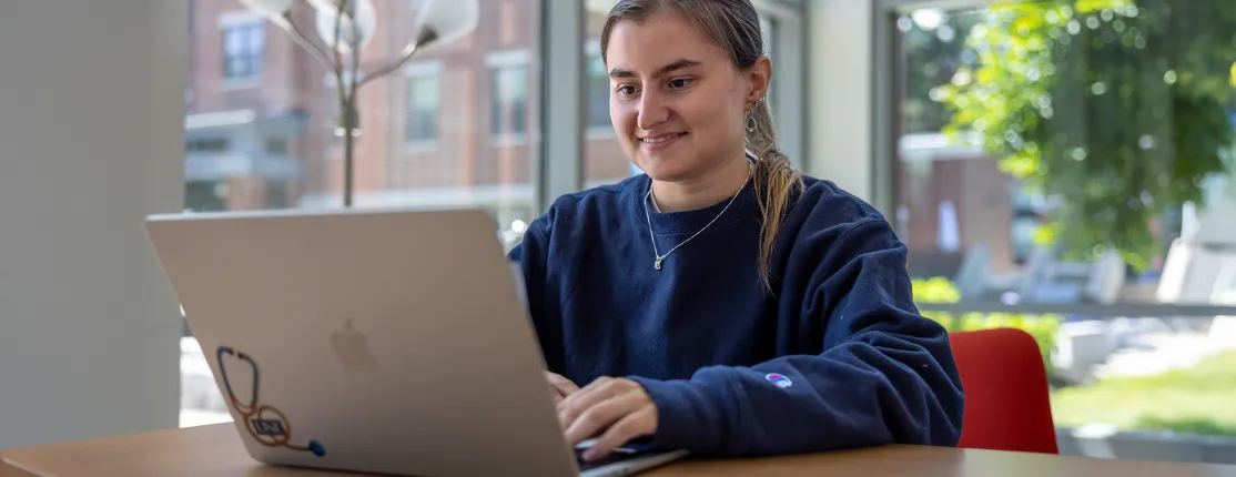 A U N E student works on their laptop in a bright campus building with large windows overlooking campus grounds.