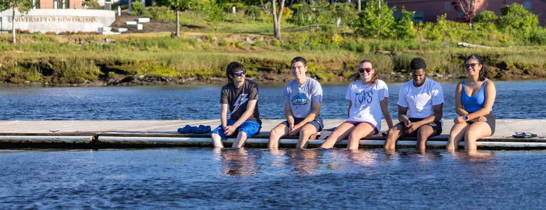 Five students sit side by side on a floating dock with their feet dangling in the water, smiling and relaxing on a sunny day. The University of New England sign and lush campus greenery are visible across the river in the background.