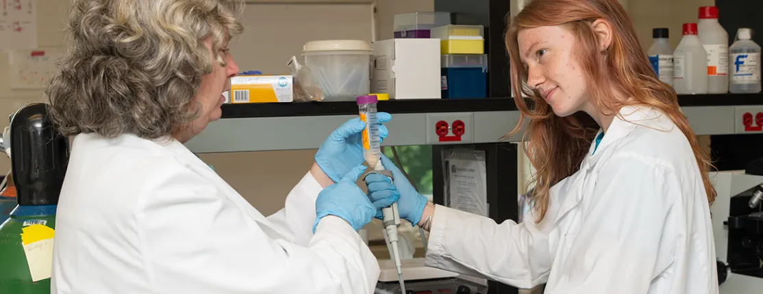 A faculty member in a lab coat demonstrates pipetting technique to a student in a research laboratory setting.