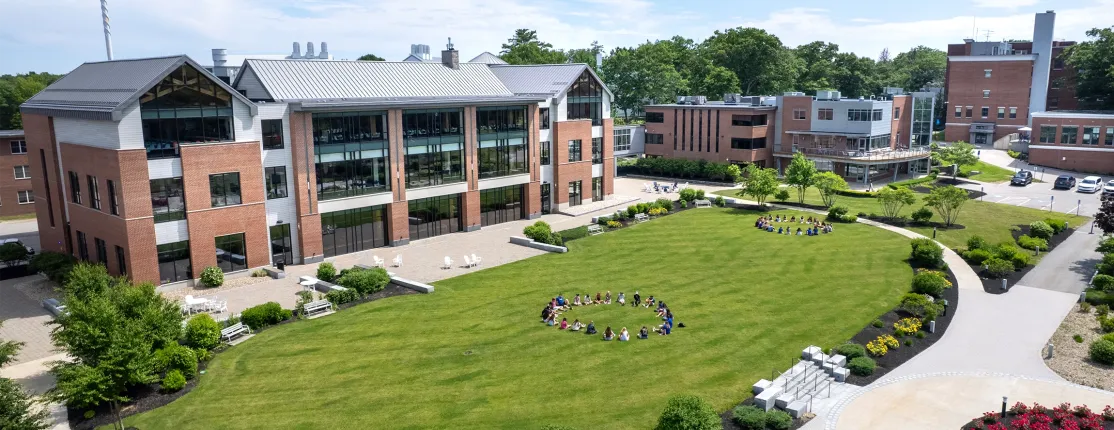 Aerial view of two groups of students sitting in circles on a green campus lawn surrounded by modern brick buildings on a sunny day.