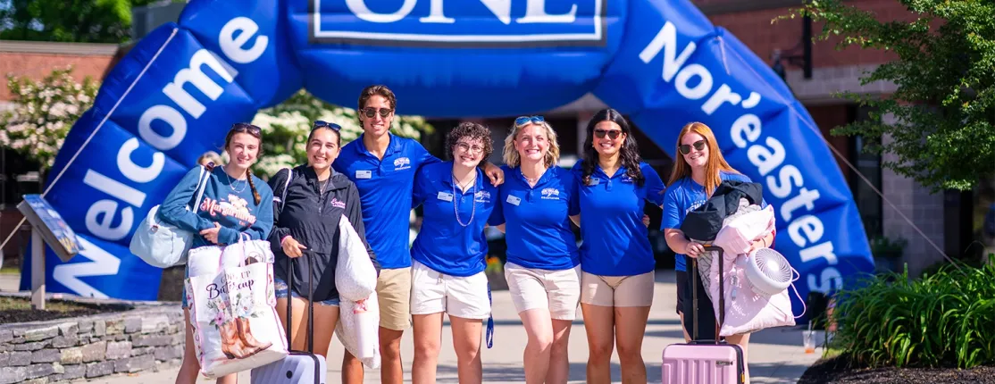 Orientation staff in blue U N E polos welcome an incoming student with luggage under a "Welcome Nor'easters" arch.