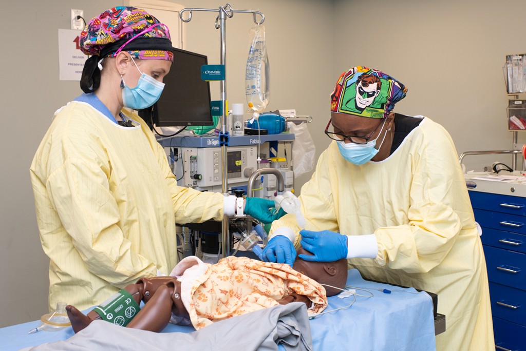 Two U N E students wearing surgical scrubs work on a patient simulator
