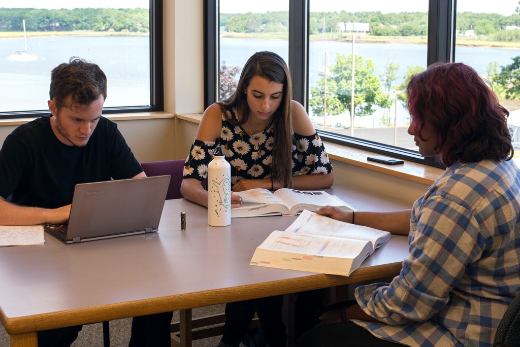 Three U N E students study together a table next to windows with a view of the ocean