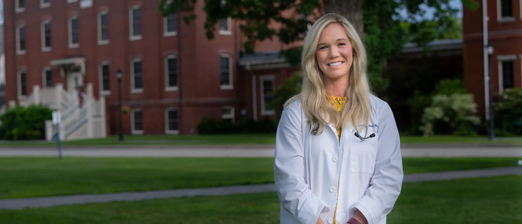 Woman stands smiling in front of camera against UNE campus green