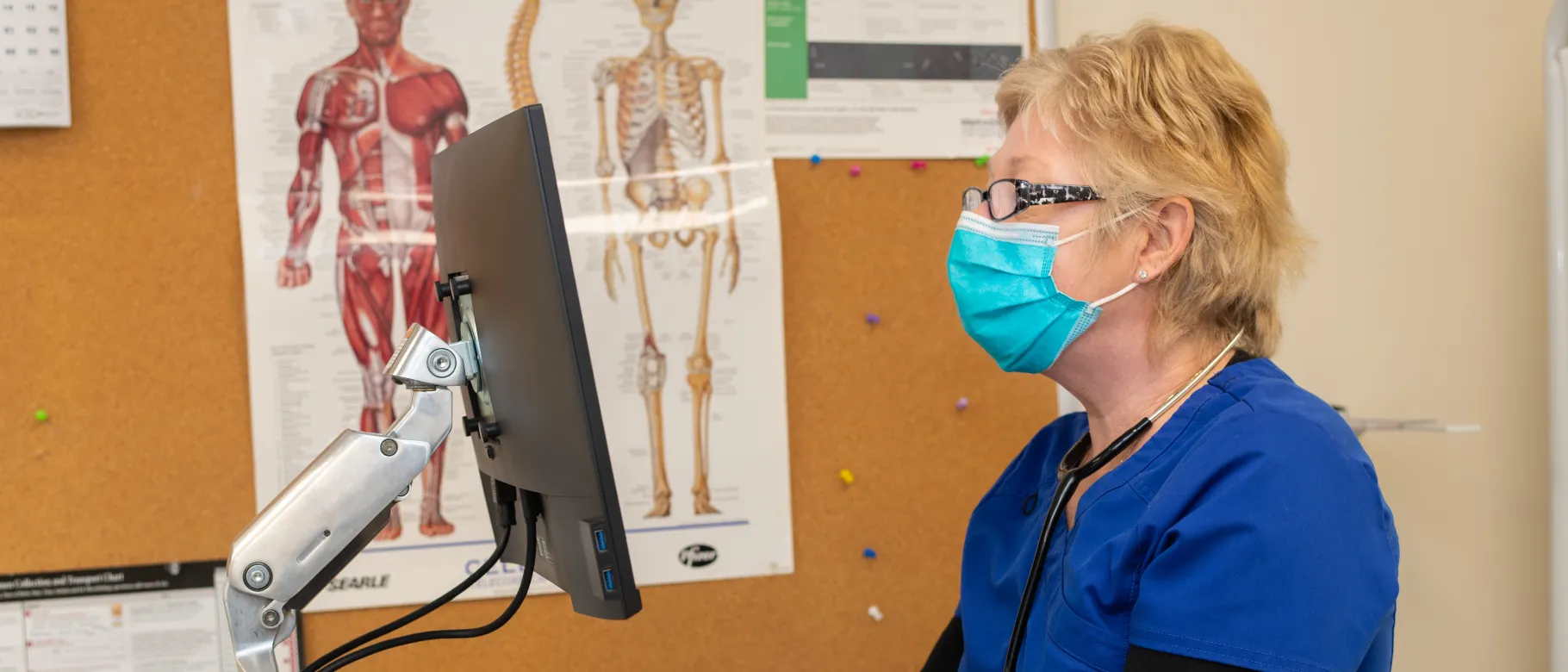 Woman with blonde hair in scrubs stands at computer in campus health center
