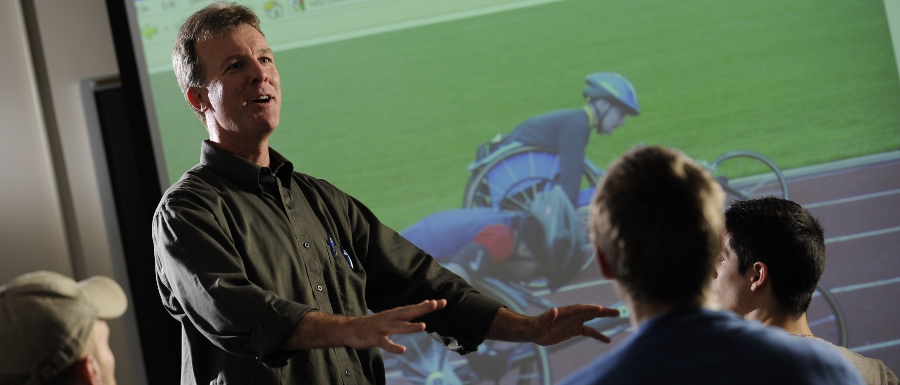 UNE Professor Jim Cavanaugh teaches students in front of a screen with cyclists in view
