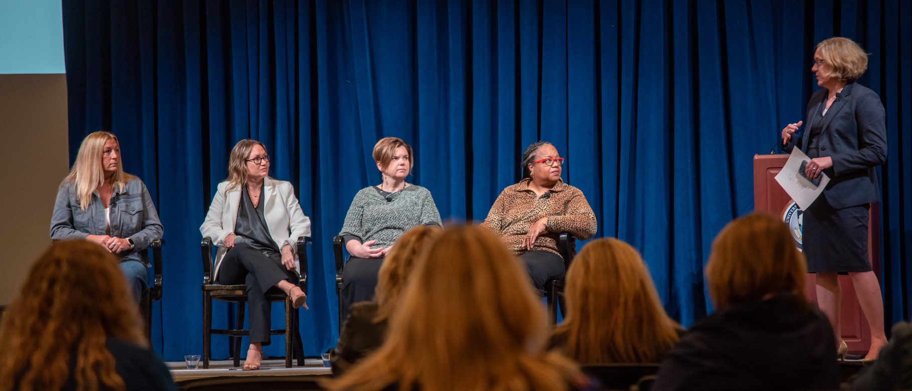 Photo shows five women on stage, four sit in chairs and another stands to moderate a panel discussion