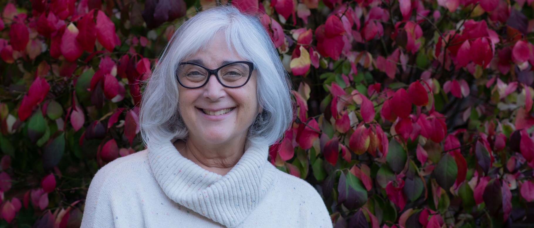 Photo of Shelley Cohen Konrad in white against a background of red leaves. 