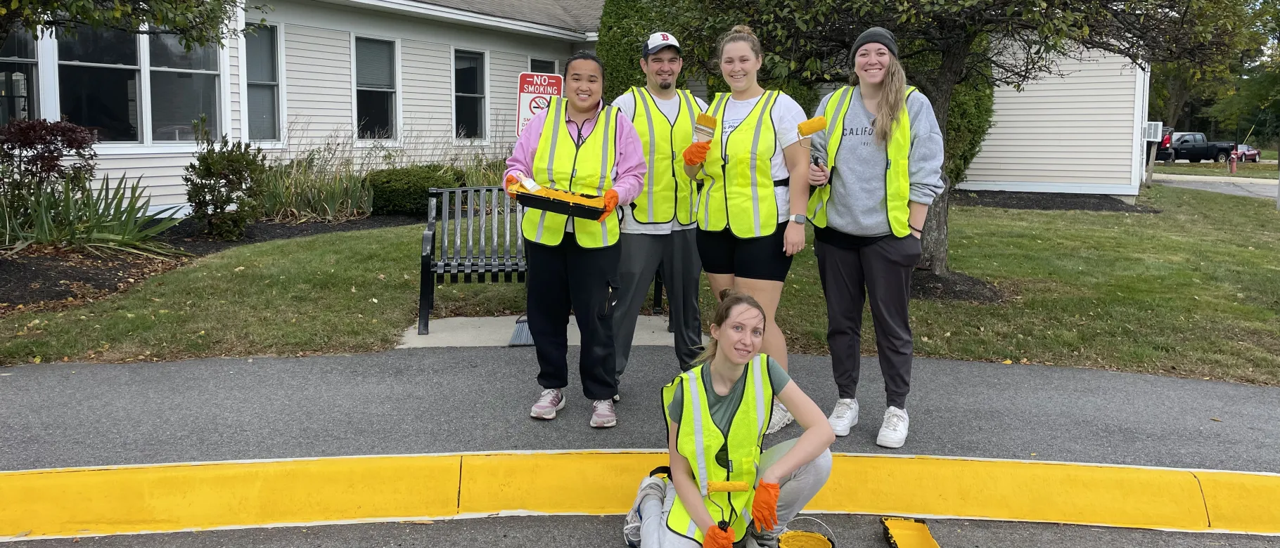 A group of students wearing yellow safety vests pose in front of the Westbrook Housing building displaying the painted sidewalk curbs they completed to enhance safety