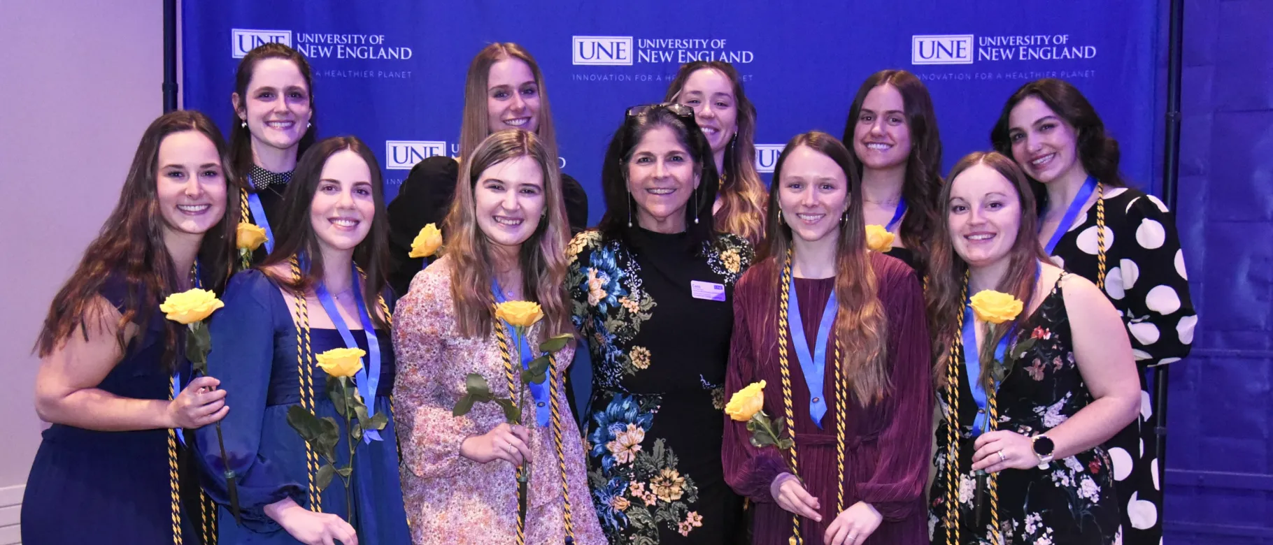 A group of students and faculty pose for a photo against a UNE-branded step-and-repeat
