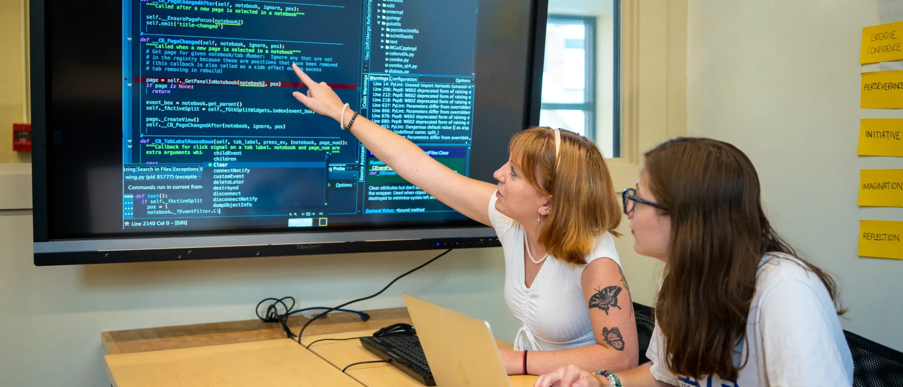 Two female students sit in front of a computer with large numbers of data appearing on a TV screen