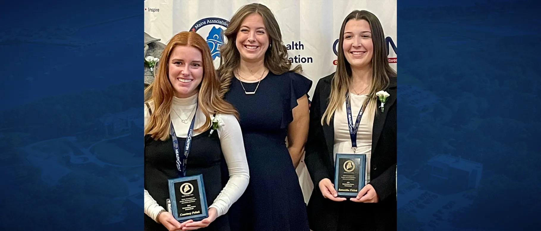 Two female students pose for a photo with a third woman while holding award plaques